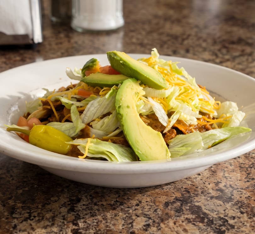 Taco salad with chili, lettuce, cheese, avocado, tomatoes, and a yellow pepper in a white bowl.