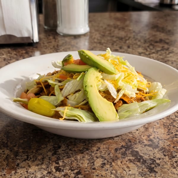 A taco salad with ground beef, lettuce, cheese, tomatoes, avocado slices, and a pepperoncini.