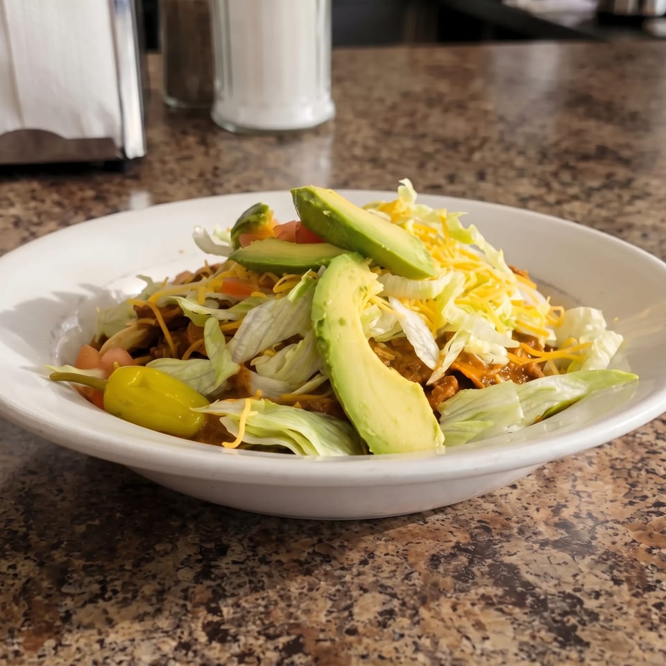 A taco salad with ground beef, lettuce, cheese, tomatoes, avocado slices, and a pepperoncini.